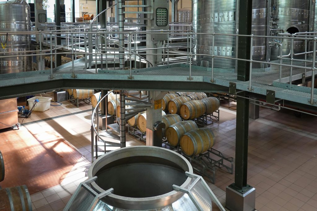 Interior view of a modern wine cellar featuring wooden barrels and large steel fermentation tanks.
